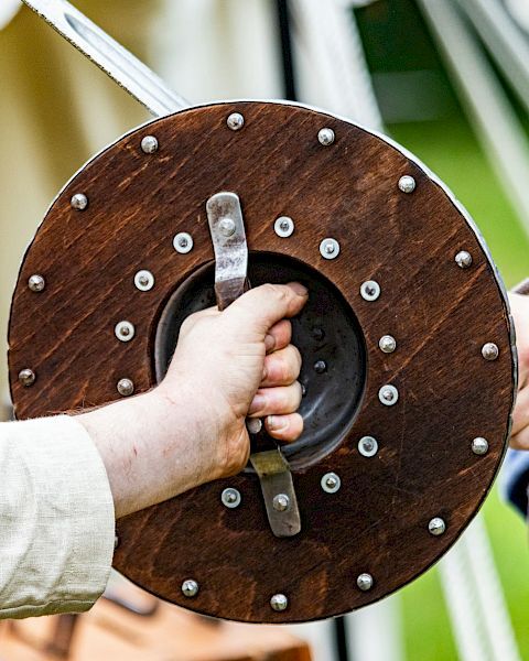 Two people, one in medieval-style clothing, are holding a wooden shield with metal rims during a historical reenactment or event.