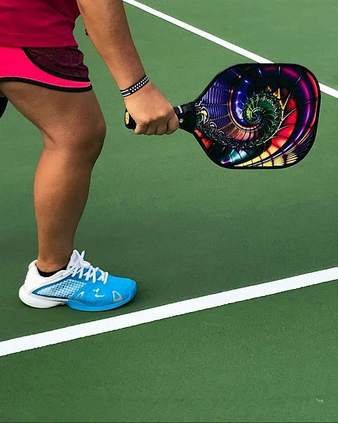 A person in sports attire holding a colorful pickleball paddle on a green court, preparing to play.