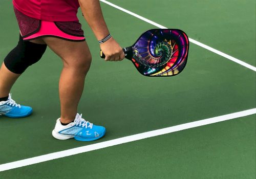 A person in sports attire holding a colorful pickleball paddle on a green court, preparing to play.