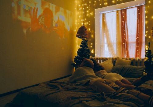 A cozy room with string lights, a projected movie, a bed with people lying in it, a window with curtains, and small Christmas trees.