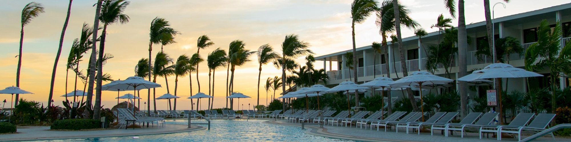 A serene resort pool area with lounge chairs, umbrellas, and tall palm trees under a sunset sky, adjacent to a building with balconies.