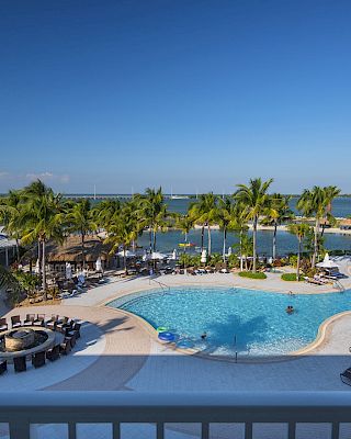 A tropical resort features a large pool surrounded by palm trees, lounge chairs, and a fire pit area, overlooking a serene body of water.