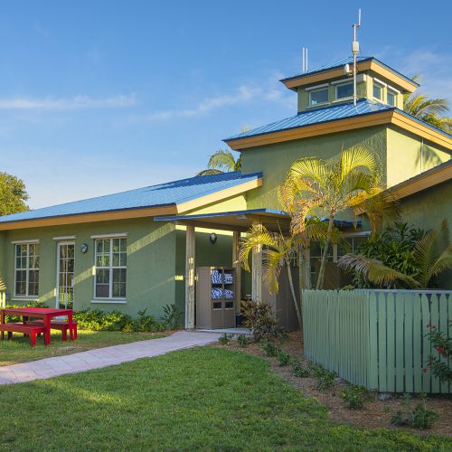 The image shows a small, green building with a blue roof, surrounded by greenery and outdoor benches on a sunny day.