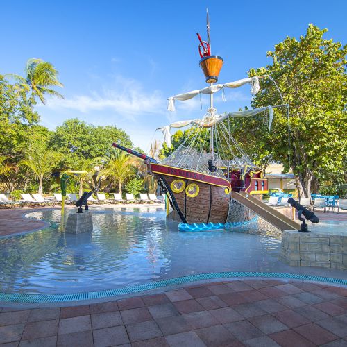 A children's splash pool with a pirate ship-themed play structure, complete with slides and water cannons, surrounded by palm trees and sun loungers.