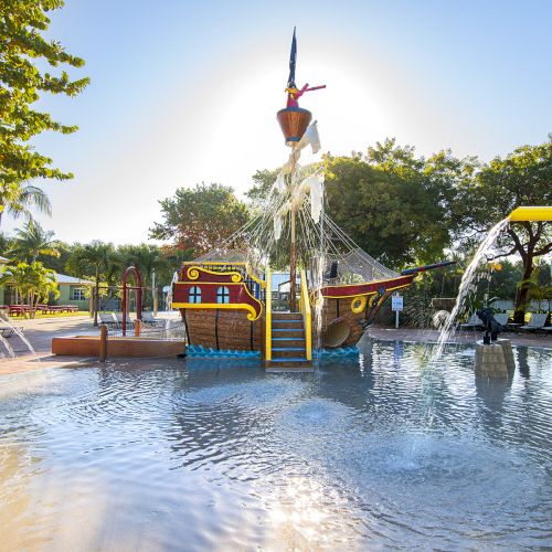 The image shows a splash pad with a pirate ship play structure, water features, and surrounding trees under a clear sunny sky.