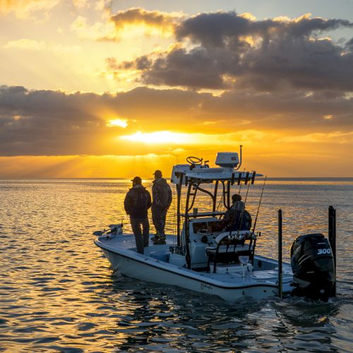 A small boat with people on board is floating on calm waters during a vibrant sunset, with the sky filled with dramatic clouds.