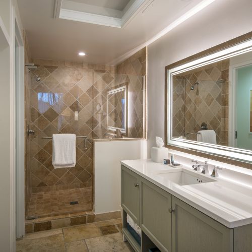 A modern bathroom with a double sink vanity, large mirror, and tiled walk-in shower with a towel hanging on the wall. Hooks are on the left wall.