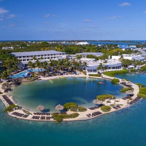 An aerial view of a resort with a lagoon, surrounded by buildings, palm trees, sun loungers, and umbrellas, set against a backdrop of blue water.