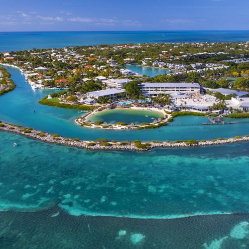 Aerial view of an island resort surrounded by turquoise waters and lush greenery, with clear skies overhead.