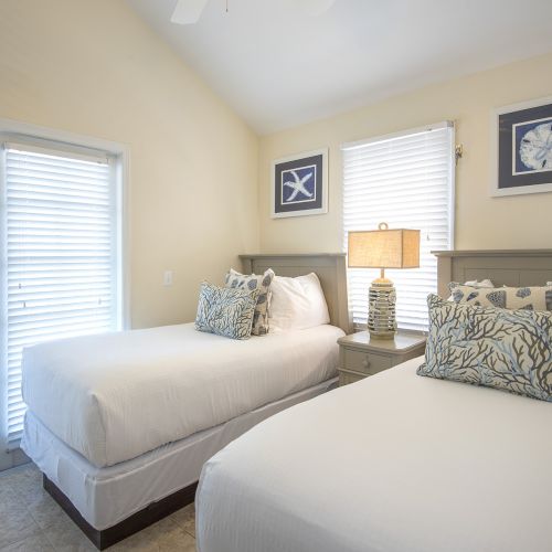 A bedroom with two single beds, white bedding, decorative pillows, a lamp on a nightstand between, and wall art above each bed.