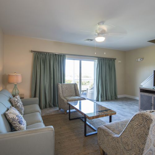 A living room with a couch, two chairs, a coffee table, TV, and a lamp near sliding doors with green curtains looking out onto a bright view.