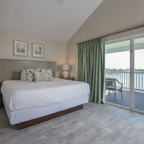 A well-lit bedroom with a large bed, side tables, and lamps. Glass doors lead to a balcony with a scenic water view outside.