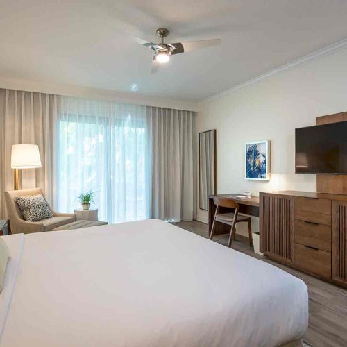 A modern bedroom with a large bed, TV, wooden furniture, a desk, and a chair. Curtains cover a window, and a ceiling fan is on.