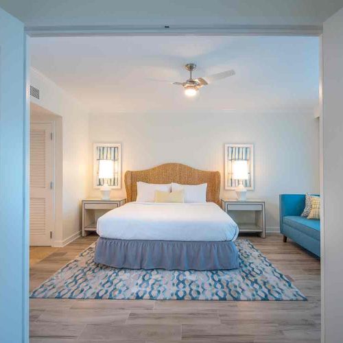 A bedroom with a large bed, two bedside tables with lamps, a ceiling fan, and a blue couch viewed through open double doors, on a patterned rug.