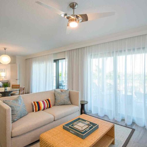 This image shows a modern living room with a beige sofa, a coffee table, a ceiling fan, and large windows with sheer curtains. A bedroom is visible in the background.