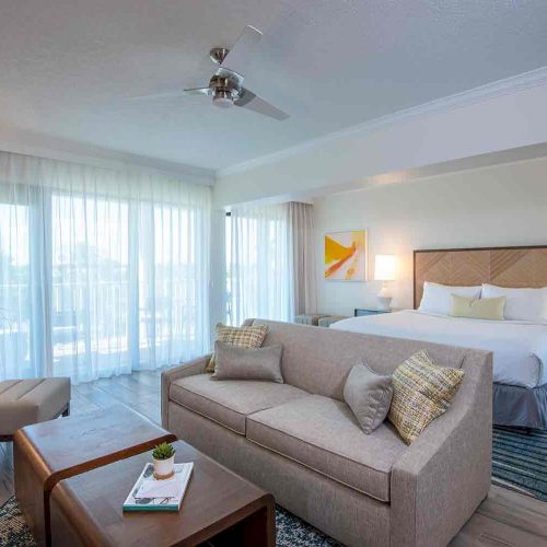 A modern hotel room featuring a bed, seating area with an armchair and sofa, coffee table with magazines, bedside tables, and large windows with sheer curtains.