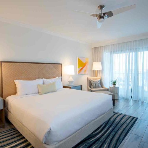 This image shows a modern bedroom with a large bed, bedside tables with lamps, a chair, a rug, and sheer curtains over glass doors.