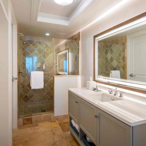 This image shows a modern bathroom with a double sink vanity, illuminated mirror, and a glass-enclosed shower with tiled walls and flooring.