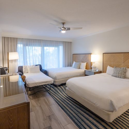 The image shows a modern hotel room with two beds, a chair, a TV on a stand, and bedside lamps. The room has a striped rug and a window.