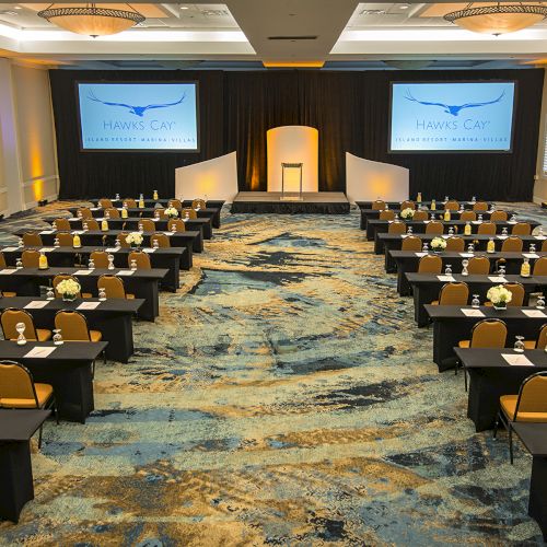 A conference room is set up with rows of tables and chairs facing a stage with a podium and twin display screens, ready for a presentation.