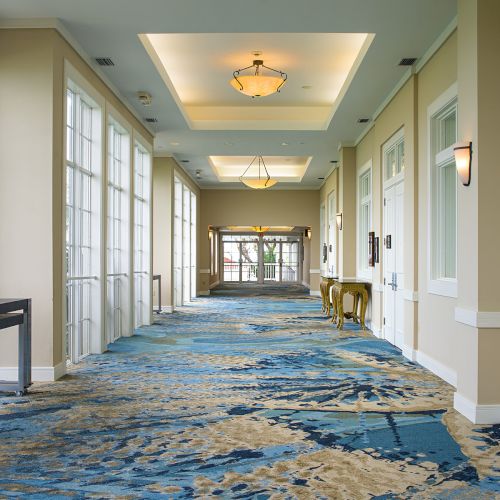 A brightly lit corridor features large windows, patterned blue and sandy carpet, and a few tables along the walls, creating an inviting atmosphere.