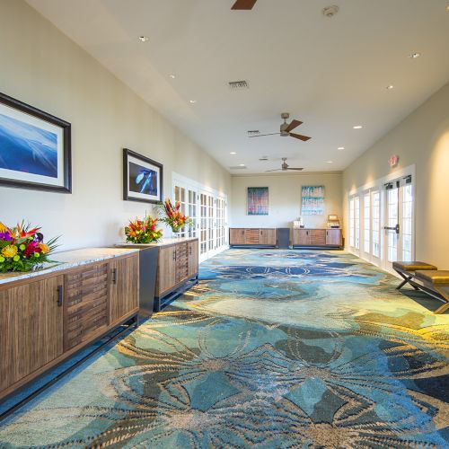 A brightly lit hallway with wooden cabinets, colorful flower arrangements, paintings on the wall, patterned blue carpet, and bench seating by the windows.