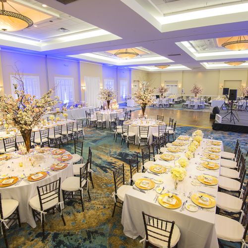 The image depicts an elegantly decorated banquet hall with round and long tables set for a formal event, featuring floral centerpieces and stage.