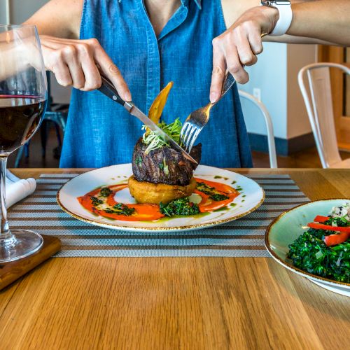 A person is cutting into a steak served with vegetables and sauce, accompanied by a glass of red wine and a side salad.