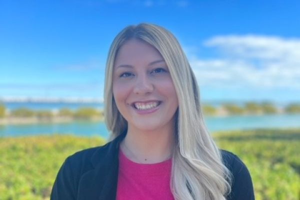 A woman with long blonde hair is standing outdoors, smiling, wearing a pink top and black jacket, with a scenic landscape and sky behind her.