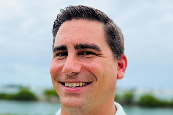 A smiling man in a light green shirt stands outdoors with greenery and a body of water in the background.