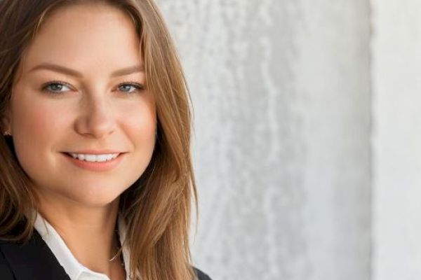 The image shows a smiling woman with long brown hair wearing a black blazer and a white shirt, standing in front of a textured, light-colored wall.