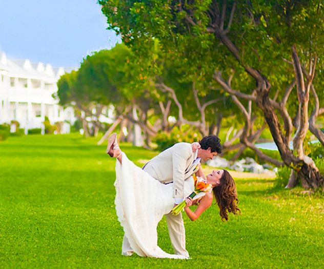 A couple in formal attire dances on a lush green lawn with white houses and trees in the background.