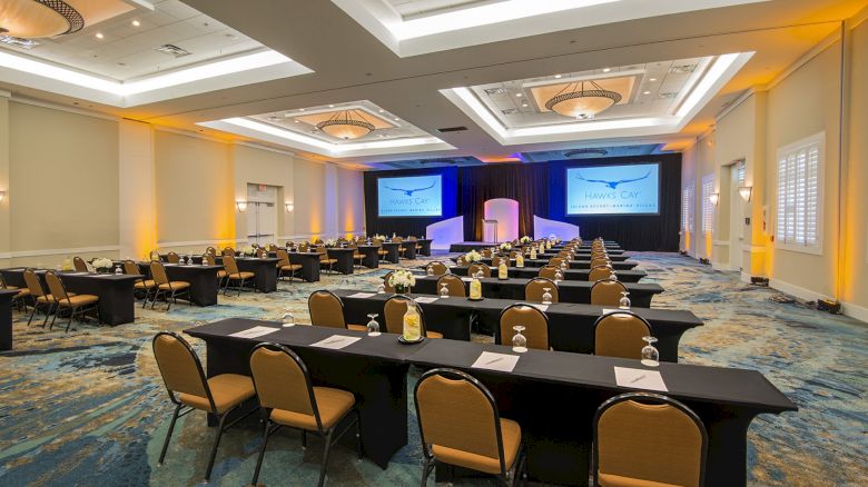 A conference room featuring rows of tables and chairs, with bottled water and notepads, and two large screens at the front displaying a presentation.