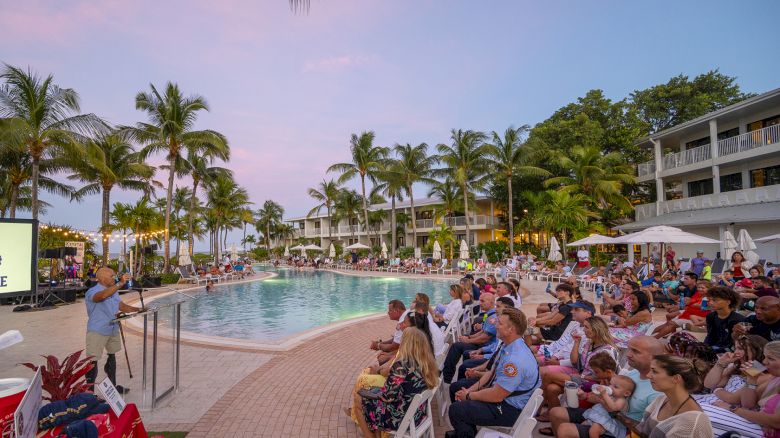 An outdoor gathering beside a pool at sunset, with people seated near a speaker. Tropical resort ambiance with palm trees and buildings.