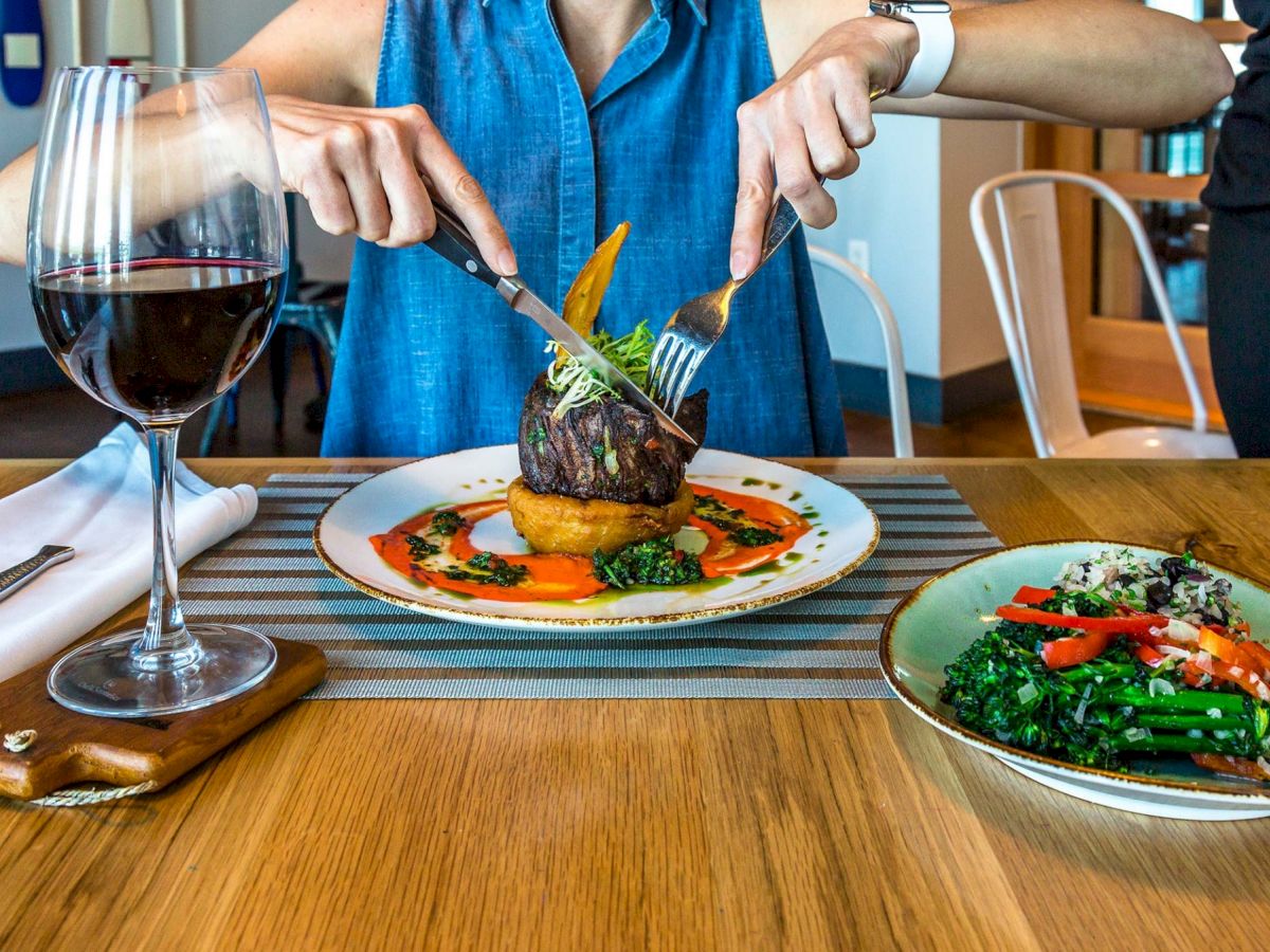 A person is cutting into a steak with a knife and fork at a restaurant, with a glass of red wine and a side dish of vegetables.