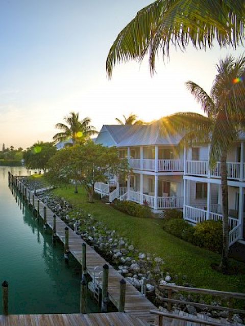 A serene waterfront scene featuring calm waters, houses with balconies, a boardwalk, and lush greenery beneath a clear sky with scattered clouds.