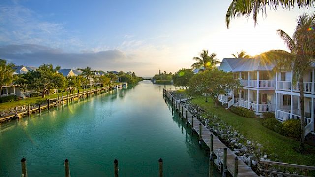 A serene waterfront scene featuring calm waters, houses with balconies, a boardwalk, and lush greenery beneath a clear sky with scattered clouds.