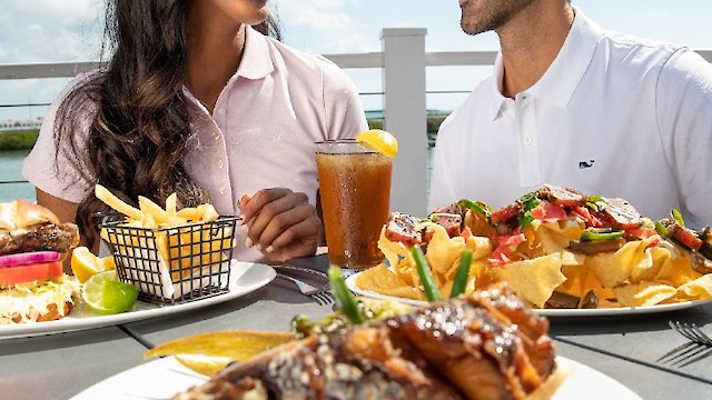 A couple is enjoying a sunny outdoor meal with dishes including nachos, fries, and fish, accompanied by a drink, all while smiling.