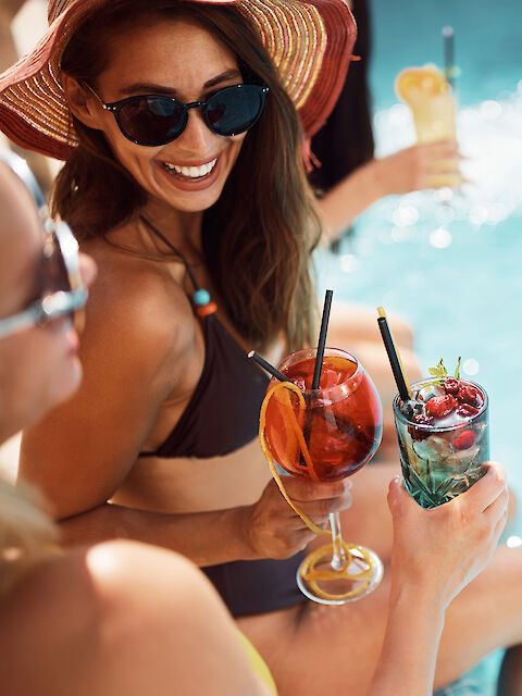 Two people sit poolside in swimsuits, holding colorful drinks with fruit garnishes. The sun is shining, creating a relaxed summer atmosphere.