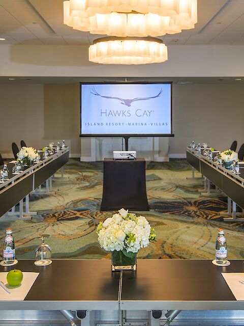 This image shows a conference room set up with U-shaped tables, chairs, water bottles, apples, and flowers, facing a screen displaying 