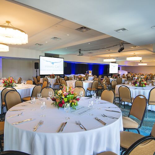 The image shows a decorated event hall with round banquet tables, floral centerpieces, chairs, and presentation screens.