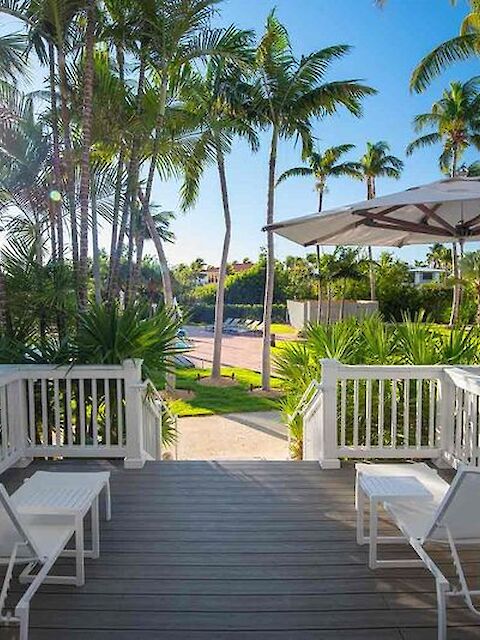 The image shows a sunlit deck with white chairs and umbrellas under palm trees, leading to a path surrounded by lush greenery and a beautiful distant view.