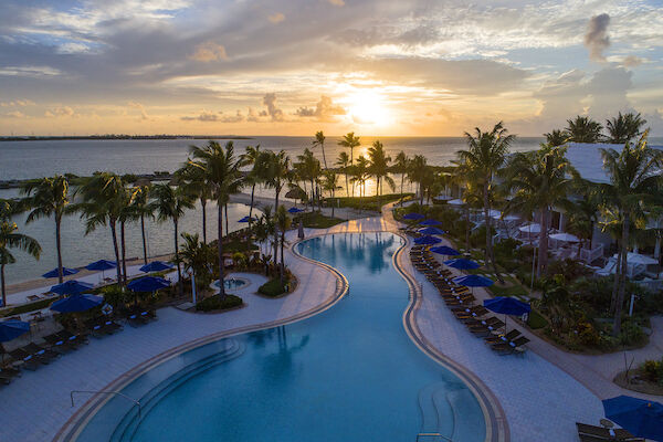 A stunning resort pool with palm trees, surrounded by lounge chairs, overlooks the ocean at sunset, creating a tropical paradise scene.