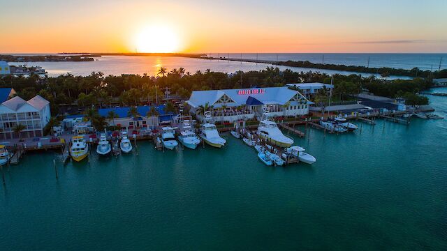 An aerial view of a marina at sunset with boats docked alongside buildings, surrounded by water and lush greenery.