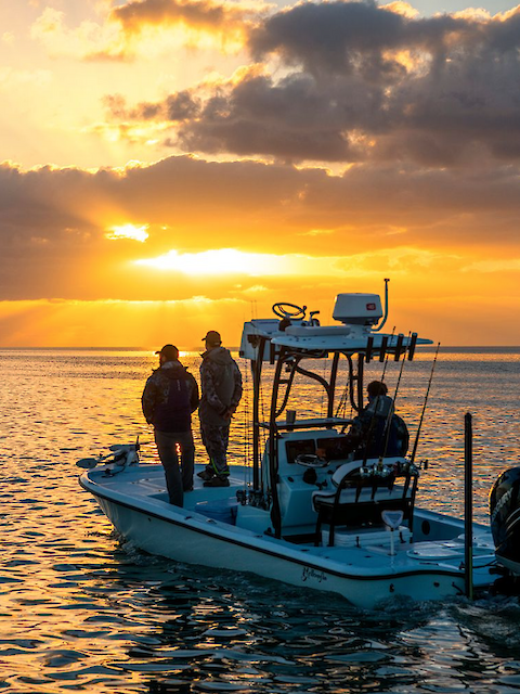 A small boat with three people is on a calm sea at sunset, with clouds and sun rays in the background.