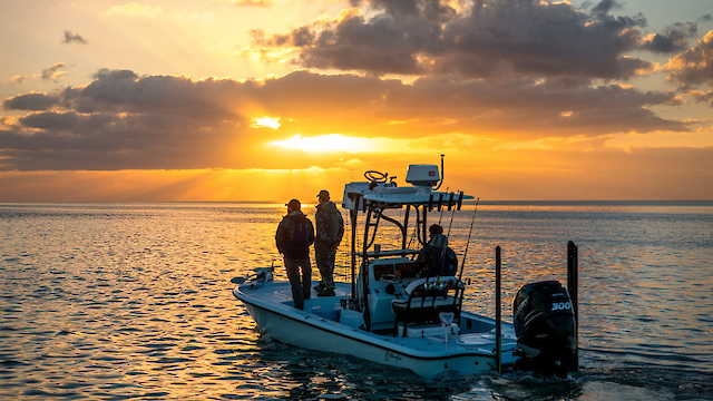 A small boat with three people is on a calm sea at sunset, with clouds and sun rays in the background.