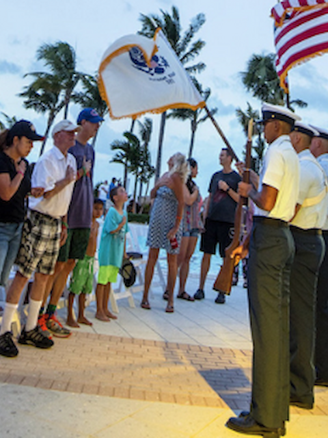 A group of people stand and observe a color guard holding flags near a poolside area with palm trees, in what appears to be a ceremony.