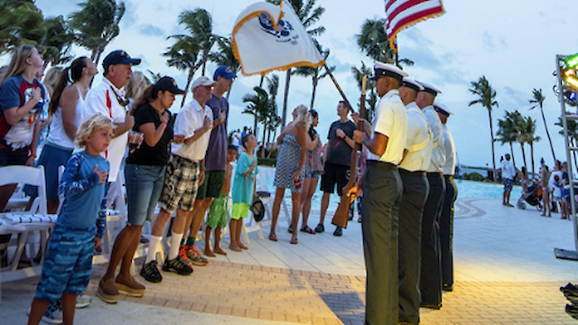 A group of people stand and observe a color guard holding flags near a poolside area with palm trees, in what appears to be a ceremony.