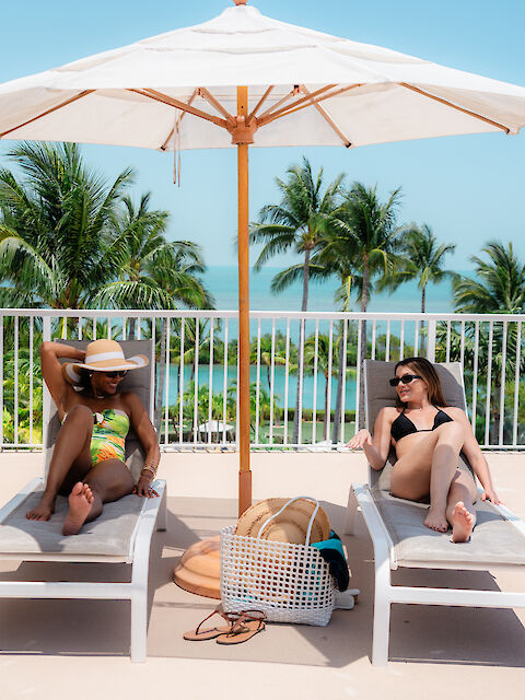 Two people relaxing on lounge chairs under a sun umbrella, with palm trees and ocean in the background.