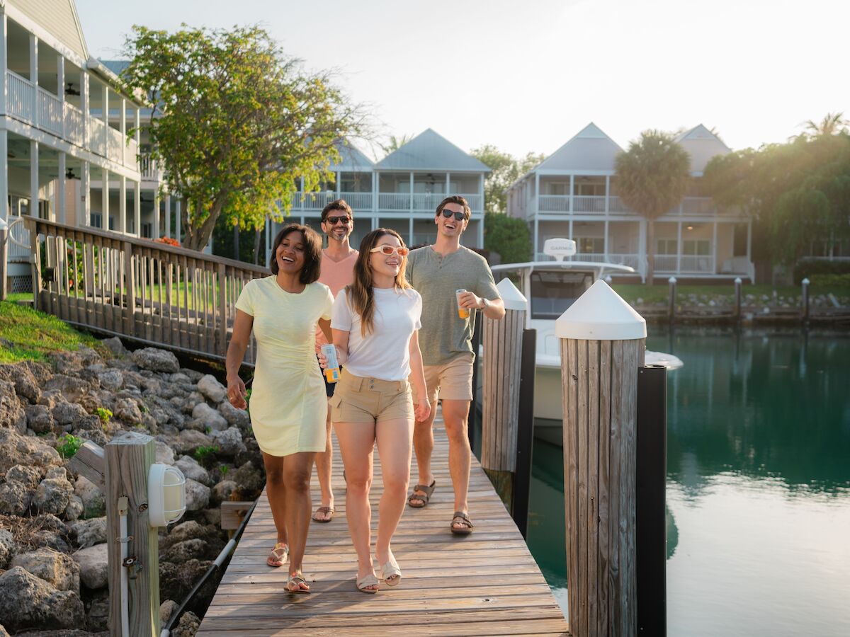 Four people are walking and laughing on a dock next to water, with houses and trees in the background. It’s a sunny day.